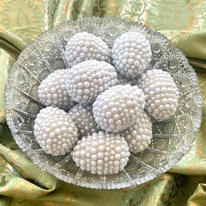 May include: A clear, cut-glass serving dish filled with white, oval-shaped objects covered in small, round, pearl-like beads. The dish sits on a green and gold patterned fabric.