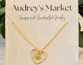 May include: A gold-colored necklace with a heart-shaped pendant. The pendant has a white background with a small, dried flower design. The necklace is displayed on a white card with the text "Audrey's Market Unique and Handcrafted Jewelry."