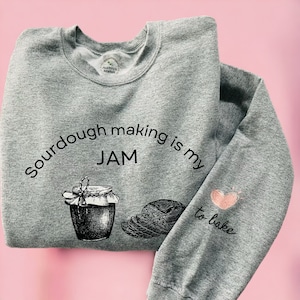Może przedstawiać: Szara bluza z nadrukiem "Sourdough making is my JAM" i rysunkiem słoika z dżemem i kromkami chleba. Na prawym rękawie znajduje się napis "to bake" i różowe serce.