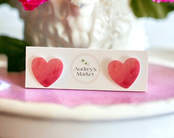 May include: A pair of pink heart-shaped earrings on a white card with the text "Audrey's Market".