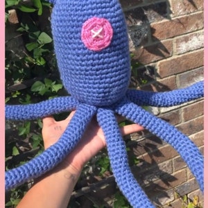 May include: A blue crocheted octopus with a pink button for an eye. The octopus is being held by a hand.
