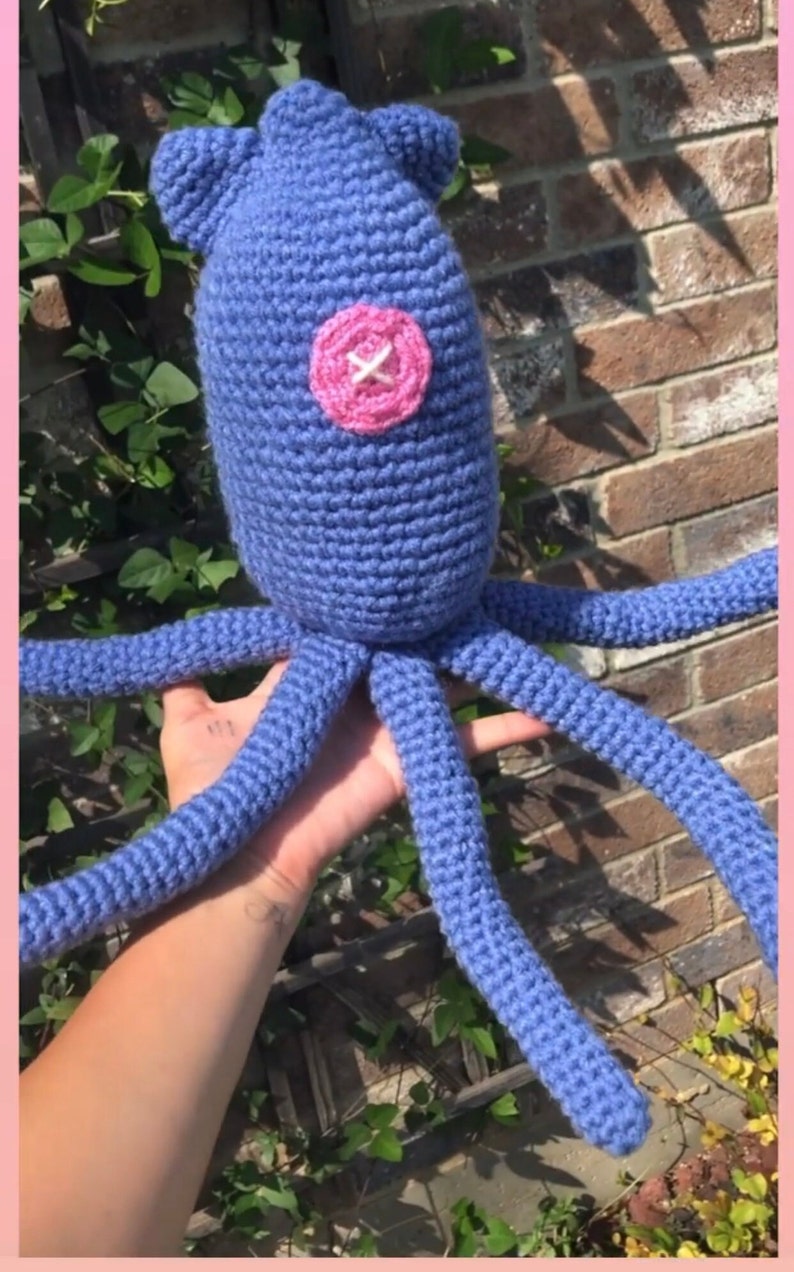 May include: A blue crocheted octopus with a pink button for an eye. The octopus is being held by a hand.