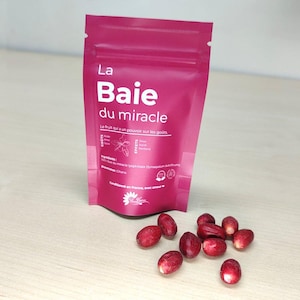 Könnte beinhalten: Ein rosa Beutel mit dem Text "La Baie du miracle" und einem weißen Blumenlogo. Der Beutel liegt auf einer hellbraunen Holzoberfläche, vor ihm liegen kleine rote Beeren verstreut.