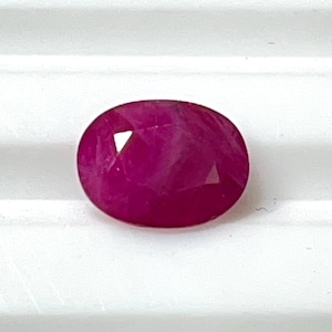 May include: A faceted, oval-shaped ruby gemstone. The gem is a deep, vibrant red colour, with visible internal facets that catch the light. The ruby is set against a white background.
