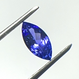 May include: A marquise-cut, deep blue gemstone is held by silver tweezers against a white background. The gemstone's facets reflect light, showcasing its brilliant color and clarity. The tweezers provide a sense of scale and context.