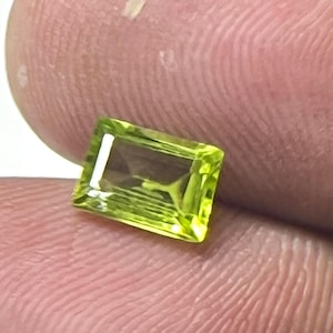 Könnte beinhalten: Ein rechteckiger, hellgrüner Edelstein mit facettierter Oberfläche. Der Edelstein wird zwischen zwei Fingern gehalten, wodurch seine Klarheit und Farbe zur Geltung kommen. Der Edelstein scheint ein Peridot zu sein, eine beliebte Wahl für Schmuck.