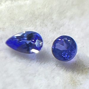May include: Two brilliant blue gemstones. One is pear-shaped, the other is round. Both stones have facets that catch the light, creating a sparkling effect. The gems are set against a soft, white background, highlighting their color and clarity.