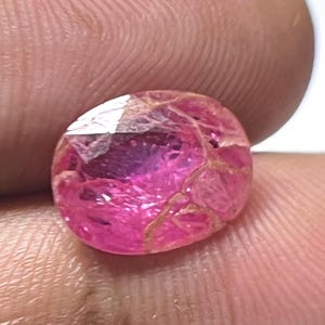 Puede incluir: Una gema facetada de forma ovalada con un vibrante tono rosa. La gema tiene inclusiones internas de color dorado y una superficie pulida. La gema se sostiene entre dos dedos sobre un fondo blanco.