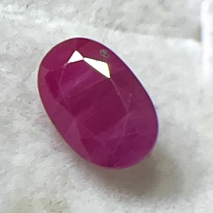 Pode incluir: Uma gema de rubi facetada em forma oval. O rubi é de cor vermelha profunda e saturada com múltiplas facetas que refletem a luz. A gema está sobre um fundo branco.