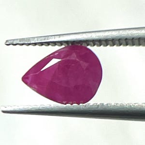 May include: A faceted, pear-shaped ruby gemstone held by tweezers. The ruby is a deep, saturated pink-red color, with visible internal reflections. The tweezers are silver-colored and hold the gemstone against a plain white background.