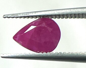 Puede incluir: Una gema de rubí facetada en forma de pera, sujeta con pinzas. El rubí es de un color rosa-rojo intenso y saturado, con reflejos internos visibles. Las pinzas son de color plateado, sobre un fondo blanco.