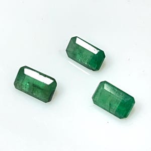 May include: Three rectangular emerald gemstones with a vibrant green hue are displayed against a white background. Each gem has a faceted cut, reflecting light and showcasing its clarity. The gems are arranged in a scattered pattern.