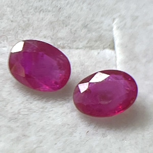 Może przedstawiać: Dwa owalne, żywe czerwone rubiny. Kamienie szlachetne są prezentowane na miękkim, białym tle, ukazując ich wypolerowane powierzchnie i wewnętrzny blask. Rubiny mają głęboki, bogaty odcień czerwieni.