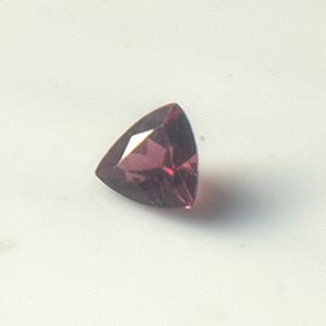 Puede incluir: Una gema triangular facetada de un profundo tono púrpura rojizo. La gema se encuentra sobre un fondo blanco, mostrando sus superficies pulidas y bordes afilados. La forma de la gema es un triángulo equilátero perfecto.