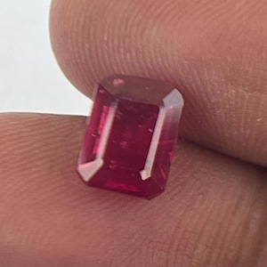 May include: A faceted, rectangular gemstone with a deep red hue. The gem has a polished surface and appears to be held between fingers. The cut of the stone reveals multiple facets, reflecting light and adding to its brilliance.
