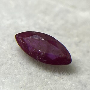 Puede incluir: Una gema roja oscura con talla marquesa y facetas. La gema es de un color rojo oscuro intenso, con una superficie pulida que refleja la luz. La forma es alargada y puntiaguda en ambos extremos, típica de un corte marquesa. Es probable que sea un rubí.