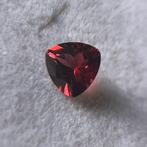 Puede incluir: Una gema triangular facetada de color rojo intenso, posiblemente un granate, descansa en una caja de joyería blanca texturizada. Las facetas de la gema capturan la luz, mostrando un color rico y oscuro. La presentación general sugiere joyería fina o gemas.