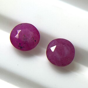 Puede incluir: Dos rubíes redondos facetados de un tono magenta rosado intenso. Cada piedra preciosa presenta una superficie pulida con inclusiones naturales visibles y un corte de múltiples facetas que refleja la luz, mostrando un color vibrante y saturado.