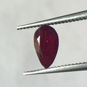 Könnte beinhalten: Ein facettierter, birnenförmiger Rubin, der von einer silbernen Pinzette gehalten wird. Der Rubin hat eine tiefe, satte rote Farbe mit sichtbaren Facetten, die das Licht einfangen. Die Pinzette befindet sich vor einem schlichten, weißen Hintergrund.