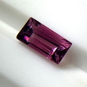 May include: A rectangular, baguette-cut gemstone with a deep purple and magenta hue. The stone features faceted surfaces that reflect light, creating variations in color intensity across its elongated, geometric shape and polished, translucent finish.
