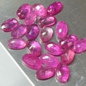 Puede incluir: Una colección de gemas rosas facetadas de corte ovalado. Las gemas son de un color magenta vibrante y parecen estar dispersas sobre una superficie metálica. Las piedras tienen una calidad pulida y reflectante.