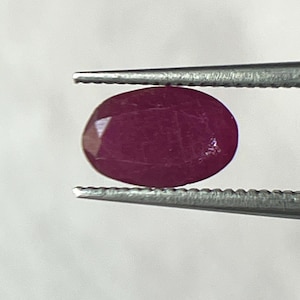 May include: A faceted, oval-shaped ruby gemstone held by tweezers. The ruby is a deep, rich red color, with visible internal inclusions. The tweezers are silver-colored and provide a contrasting background.