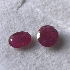 Puede incluir: Dos gemas de rubí ovaladas y facetadas. Los rubíes son de color rojo intenso y están sobre un fondo blanco texturizado. Las gemas se muestran en una caja de joyería.