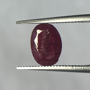 Könnte beinhalten: Ein ovaler, tiefroter Edelstein wird von einer silbernen Pinzette gehalten. Der polierte Stein weist interne Einschlüsse und einen leichten Glanz auf. Die Pinzette ist vor einem unscharfen Hintergrund scharf. Dies ist ein Edelstein.