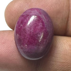 Op de afbeelding: Een gepolijste, ovaalvormige edelsteen met een diepe magentakleur. Het oppervlak van de steen heeft een glanzende glans, die het licht reflecteert. De edelsteen wordt tussen de vingers gehouden, wat de grootte en kleur laat zien.