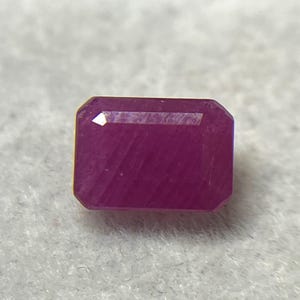 Puede incluir: Una piedra preciosa rectangular de corte esmeralda con un tono magenta rosado profundo y saturado. La piedra presenta estrías internas paralelas distintivas y bordes facetados que captan la luz, mostrando una calidad translúcida y una superficie pulida.