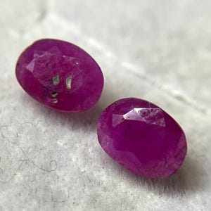 Könnte beinhalten: Zwei oval geschliffene, tief magentafarbene Rubine mit facettierten Oberflächen. Die Edelsteine weisen natürliche Einschlüsse und eine polierte Oberfläche auf, die das Licht auf ihren facettierten Oberseiten reflektiert.
