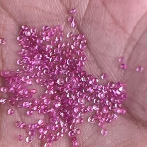 May include: A collection of small, faceted pink gemstones, possibly sapphires or rubies, are held in a hand. The gems are scattered across the palm, reflecting light and appearing vibrant against the skin. The overall color palette is a soft pink.