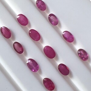 May include: A selection of oval-shaped, deep red gemstones displayed on a white surface. The gemstones are evenly spaced and have a polished, reflective appearance. The arrangement suggests a display of loose gemstones.