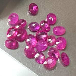 May include: A collection of oval-cut, vibrant pink gemstones. The gems are faceted, reflecting light and showcasing their clarity. The stones are scattered on a gray surface, suggesting a jeweler's workspace or display.