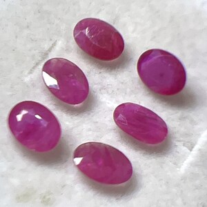 May include: Six oval-shaped, faceted rubies in varying shades of pink and red. The gemstones are displayed on a white surface, showcasing their polished surfaces and internal clarity. These rubies are suitable for jewelry making.