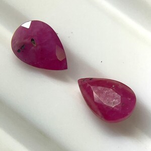 May include: Two pear-shaped, faceted rubies with a deep magenta-pink hue. Each gemstone features a polished surface with visible natural inclusions and dark internal specks, showcasing a distinct teardrop silhouette and reflective facets.