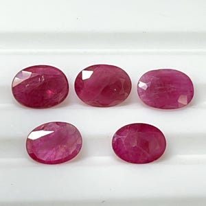 May include: Five oval-cut, faceted ruby gemstones. The rubies are a deep, rich red colour, with visible internal inclusions. The gemstones are displayed on a white surface, showcasing their brilliance and clarity. Ideal for jewellery making.