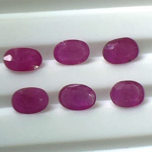 Op de afbeelding: Zes ovaal geslepen, dieprode edelstenen. De edelstenen zijn gerangschikt in twee rijen van drie, elk met een gepolijst, reflecterend oppervlak. De stenen liggen tegen een witte achtergrond, wat hun rijke kleur en vorm benadrukt. Dit zijn waarschijnlijk robijnen.