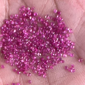Pode incluir: Uma mão segura um monte de pequenas pedras preciosas rosa. As gemas facetadas refletem a luz, criando um efeito brilhante. As gemas soltas estão espalhadas na palma da mão.