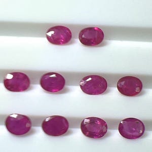 May include: An assortment of eleven oval-cut, deep red rubies. The gemstones are arranged in rows against a white background. Each ruby exhibits a faceted surface, catching the light and displaying a vibrant color.