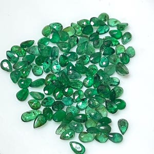 May include: A collection of emerald gemstones in various shapes and sizes, including pear and oval cuts. The gemstones are a vibrant green color and are scattered across a white surface. The gems are faceted, reflecting light.