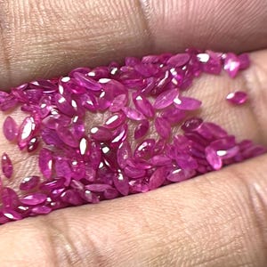 May include: A collection of small, oval-shaped, polished ruby gemstones. The gems are a vibrant pinkish-red color and are scattered across a surface. The gemstones are likely for jewelry making or collecting.