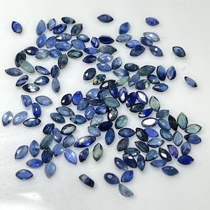 Könnte beinhalten: Eine Ansammlung von Saphiren im Marquiseschliff in verschiedenen Blautönen, von tiefem Indigo bis zu blassem Türkis. Die Edelsteine sind auf einer weißen Oberfläche verstreut und zeigen ihre polierten Facetten und natürlichen Farbvariationen. Diese Saphire eignen sich ideal für die Schmuckherstellung.