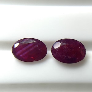 Può includere: Due pietre preziose sfaccettate di forma ovale con un colore rosso scuro profondo e saturo. Ogni pietra presenta una superficie lucida con molteplici sfaccettature geometriche che riflettono la luce, mostrando una qualità traslucida e una ricca tonalità cremisi uniforme.