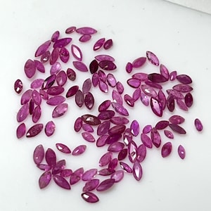 Könnte beinhalten: Eine Sammlung von Edelsteinen im Marquiseschliff in verschiedenen Rosa- und Rottönen. Die Edelsteine sind auf einer weißen Oberfläche verstreut, mit unterschiedlichen Größen und Formen. Die Edelsteine sind facettiert, reflektieren das Licht und verleihen Glanz.