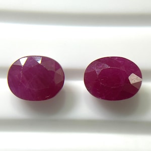 Puede incluir: Dos rubíes facetados de forma ovalada con un color rojo magenta profundo y saturado. Cada piedra preciosa presenta una superficie pulida con múltiples facetas geométricas que reflejan la luz, mostrando una calidad translúcida.