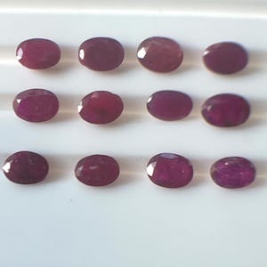 May include: An array of twelve oval-cut rubies, each displaying a deep red hue. The gemstones are arranged in three rows of four, set against a white background. The facets of the rubies catch the light, creating a sparkling effect.