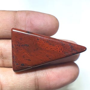 Puede incluir: Una gema triangular pulida, de color marrón rojizo intenso, que muestra patrones complejos. La superficie de la piedra es lisa y reflectante, destacando las variaciones naturales. La forma es un triángulo rectángulo, con una punta y una base plana.