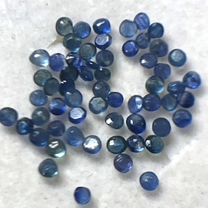 May include: A collection of small, round, faceted gemstones in shades of blue. The gems are scattered across a white surface, with variations in color from deep indigo to lighter, translucent hues. The facets catch the light, creating a sparkling effect.