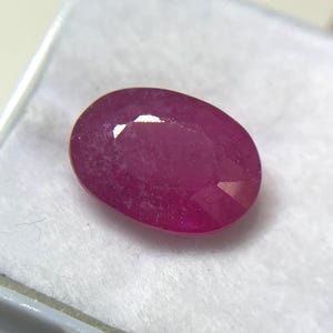 May include: An oval-cut, deep pinkish-red gemstone with a faceted surface. The stone displays a translucent quality with internal inclusions and light reflections on its polished top facets, resting on a white, textured display surface.